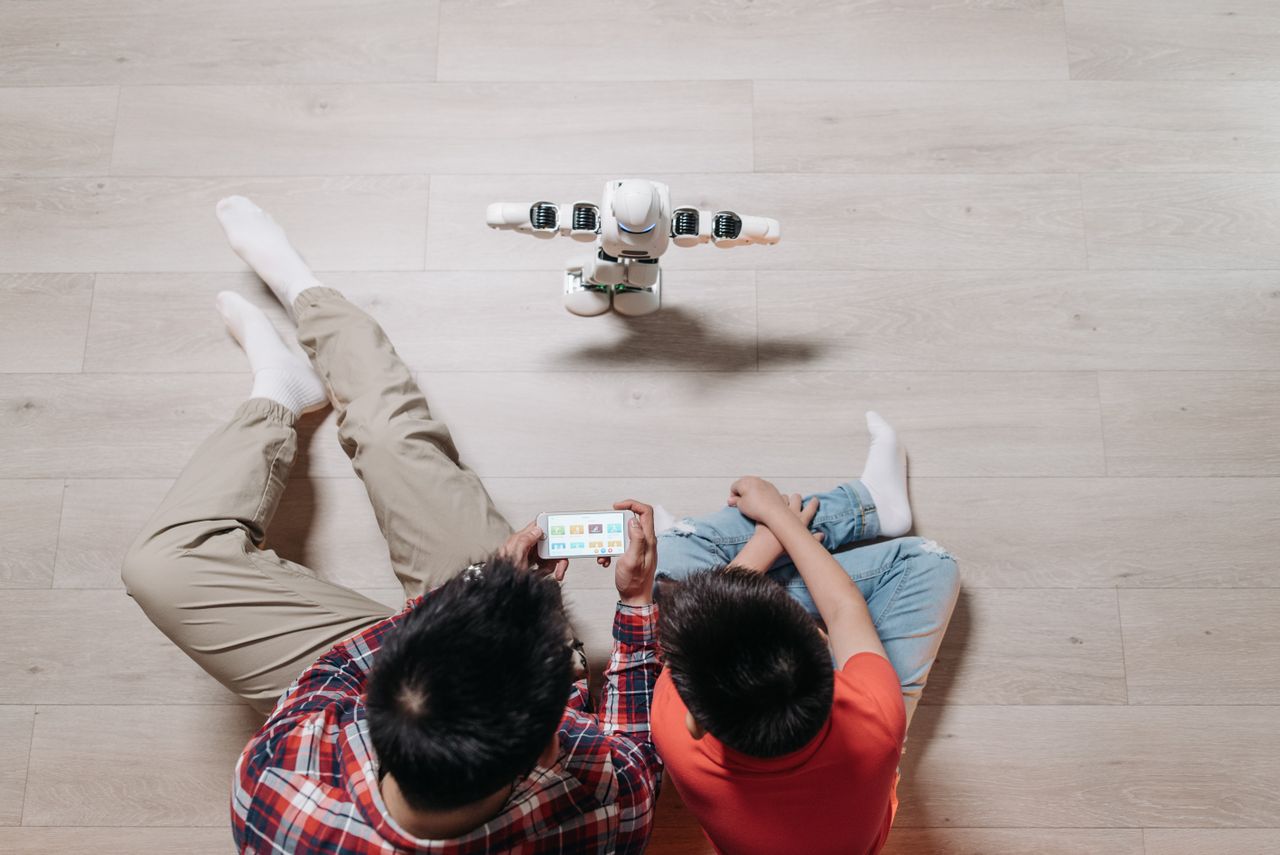 Services A father and son using a smartphone to control a small humanoid robot on wooden flooring.